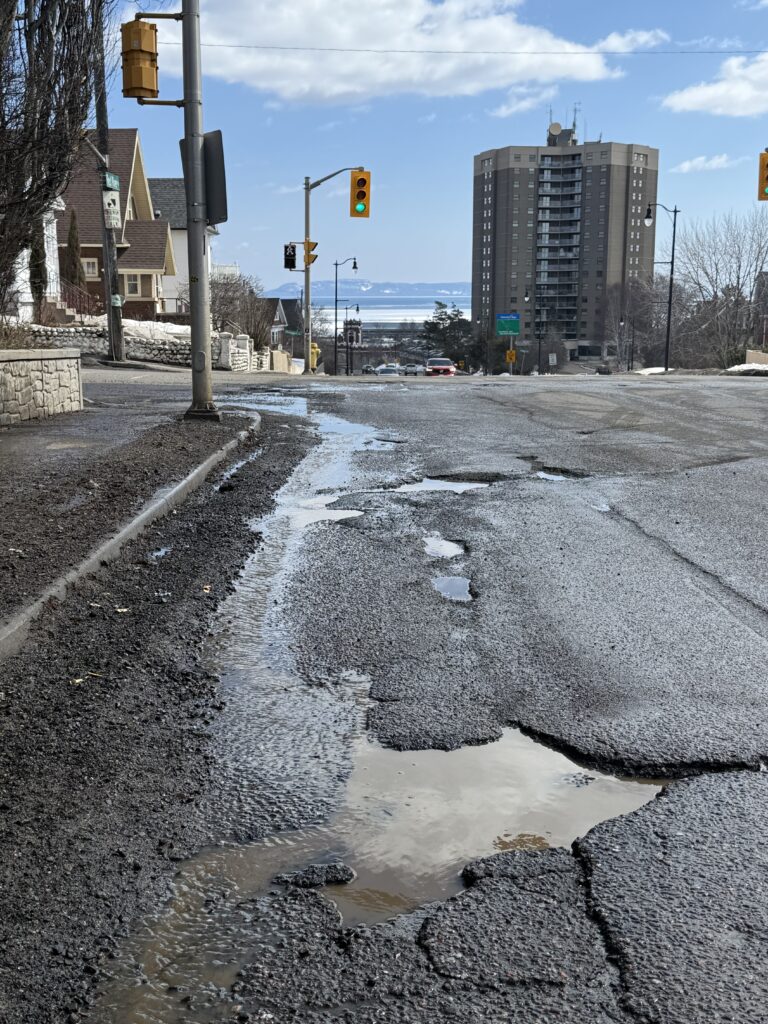 Potholes and crumbling pavement on a Thunder Bay street in spring 2026, with the Sleeping Giant visible in the background