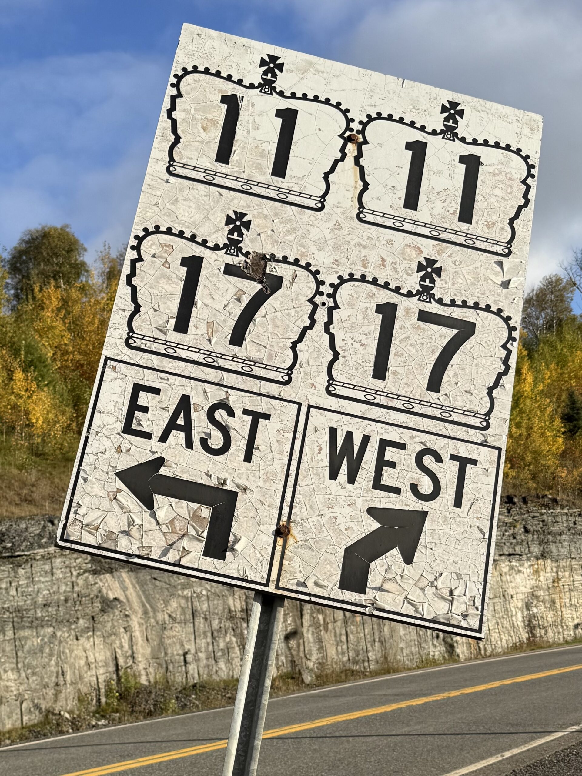 Directional sign for Highways 11 and 17 east and west near Thunder Bay, with a rock cut and autumn trees behind it.